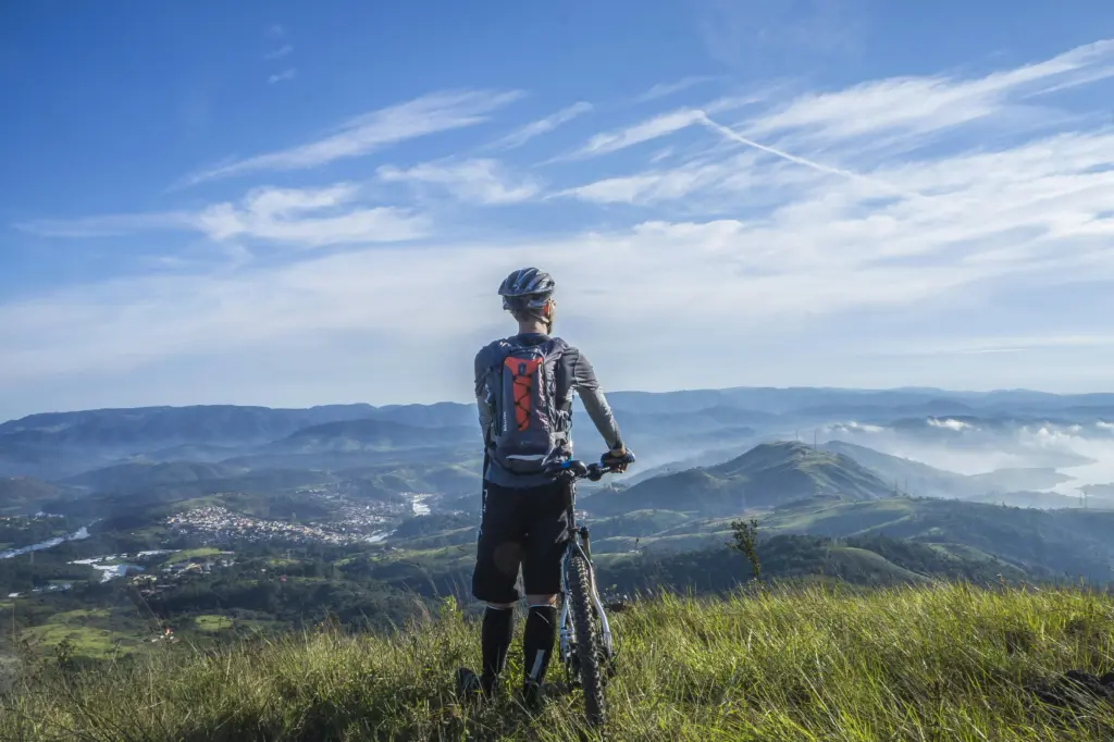Radfahrer mit Rucksack steht auf einem Berggipfel und blickt über eine grüne Hügellandschaft – aktives Sommercamping in den Bergen.