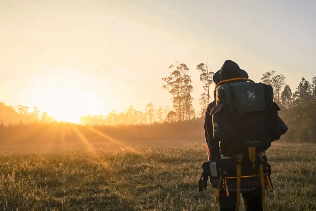 Camper mit großem Rucksack blickt beim Sommercamping in der Morgensonne über eine Wiese am Waldrand.