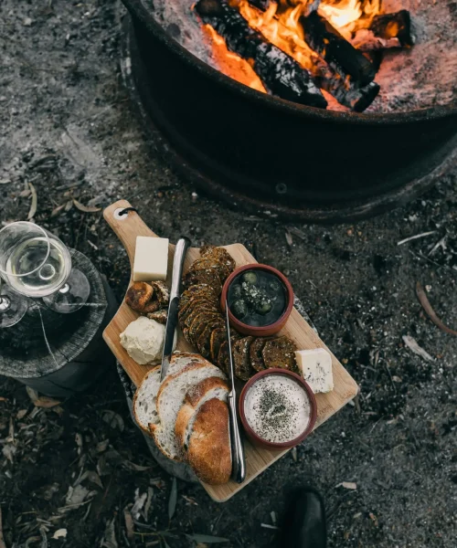 Camping Rezepte: Holzbrett mit Essen neben Lagerfeuer – rustikales Outdoor-Mahl